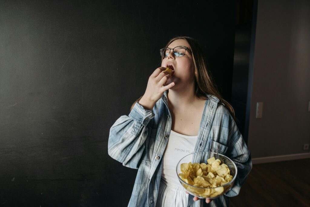 Woman eating chips indoors, illustrating emotional eating and nervous system regulation challenges in holistic fitness coaching by Pavel Danilyuk